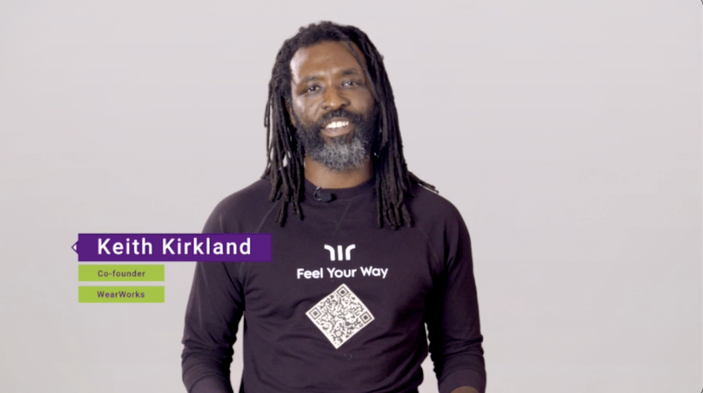 Keith Kirkland wears black longsleeved WearWorks shirt and stands in white studio setting. His title appears in purple and green box in lower-left corner 'Keith Kirkland. Co-Founder WearWorks'