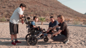 Person in Participant's cub wheelchair on a beach. Two people are in front of the chair adjusting the components while another person is leaning on the back of the chair.