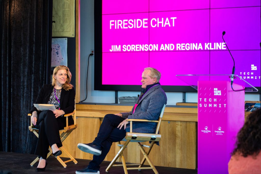 A woman wearing a black pant suit and a man wearing a checkered business jacket and dark jeans are seated on wooden chairs presenting a talk on a stage. Behind them is a pink screen that includes the text 'Fireside chat with Regina Kline and Jim Sorenson'