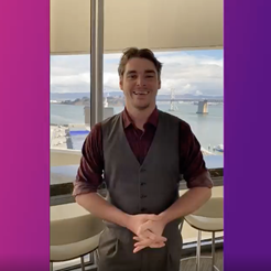RJ Mitte is standing holding his hands in front of his waist and smiling. He is wearing a collared shirt and vest and is positioned in front of a window overlooking San Francisco.
