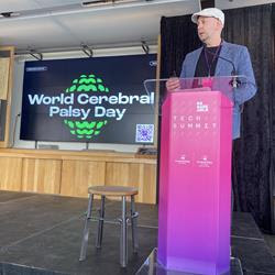 A man stands behind a lectern on a stage talking into a microphone. Behind him is a screen with the World Cerebral Palsy Day logo featured in the centre.
