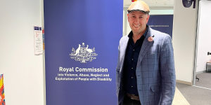 Pete Horsley is wearing a dark blue business jacket, dark pants and hat, he is smiling inside an office setting and to his right is a Disability Royal Commission branded banner.