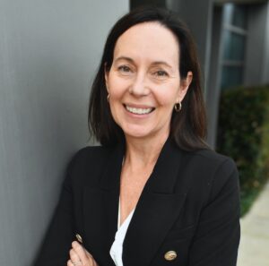Woman with dark brown hair, hazel eyes and fair skin colour. She wears a black business jacket and is smiling with her arms folded.