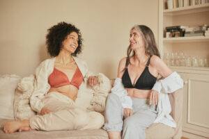 Two beautiful women wearing a Springrose front closure bra one in black and one in a burnt orange. Both women are smiling wide while seated on a lounge.
