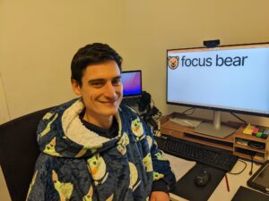 Man with dark hair and fair skin wearing a cartoon theme jumper in seated at a desk smiling with a monitor behind him that displays the focus bear logo.