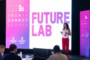 A young woman presenting on a stage in front of a large screen that says ‘Tech Summit: Future Lab’’