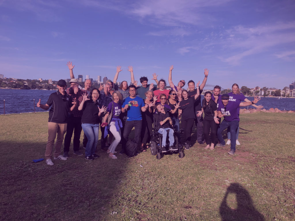 Photo of a group of diverse people with their hands in the air outside with water, the Sydney Harbour Bridge, a grass area, and blue sky in the background.