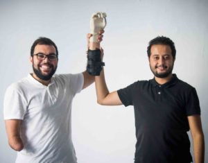 Two men standing in a studio setting both holding a bionic arm prototype.