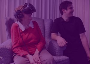 Elderly person in chair using virtual reality headset with person kneeling to their left