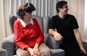 Elderly person in chair using virtual reality headset with person kneeling to their left