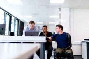 Two people standing at desk next to person in wheelchair with prosthetic arms